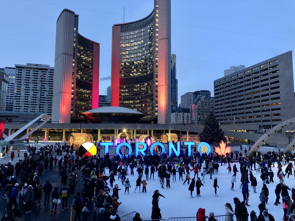 Families and visitors ice skating in Nathan Phillips Square in front of the brightly lit "TORONTO" sign at dusk. Toronto City Hall’s curved towers glow red above the busy rink creating a lively winter scene.