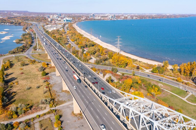 An aerial autumn view of the Burlington Skyway bridge carrying QEW traffic over Burlington Bay, with colourful fall foliage, a sandy shoreline, and the blue waters of Lake Ontario visible alongside the highway, and the Hamilton skyline in the distance.