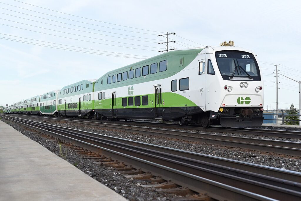 A GO Transit double-decker train in its distinctive white and green livery travelling along the tracks from Toronto to Niagara Falls, viewed from platform level.
