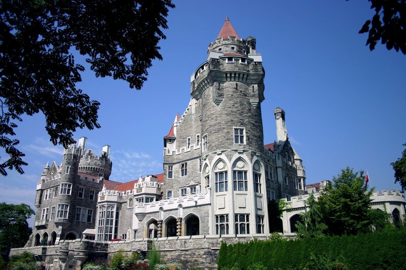 Casa Loma castle in Toronto viewed from the grounds below, showing its grey stone Gothic Revival architecture with a tall round tower topped by a red-roofed turret, arched windows, and cream-coloured lower facade, framed by lush green summer trees under a blue sky.