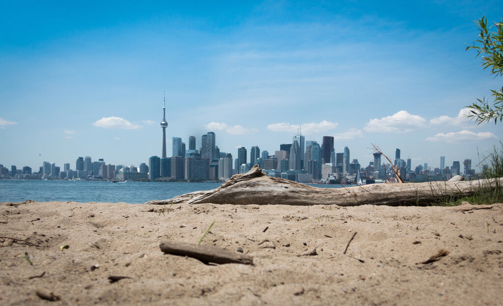 Sandy beach with driftwood in the foreground looking across Lake Ontario toward the Toronto skyline with the CN Tower rising above the city. Popular outdoor spot when visiting Toronto with kids for skyline views and lakeside space to play.