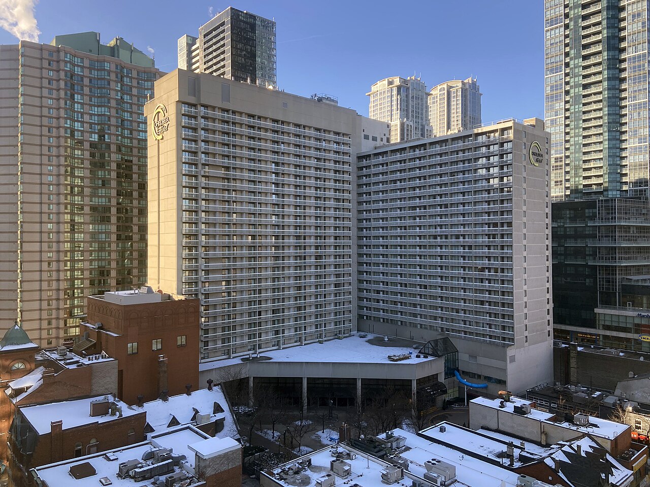 Chelsea Hotel in Toronto rises among surrounding downtown buildings, with rows of balconies and a snow covered lower rooftop, as sunlight hits the facade against a clear winter sky. It's one of the best hotels in toronto for families.