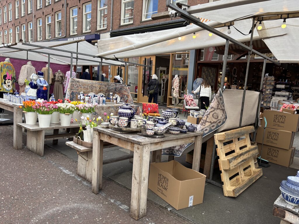 Outdoor market stall in Amsterdam displaying tulips pottery textiles and home goods on wooden tables with shoppers browsing in the background.