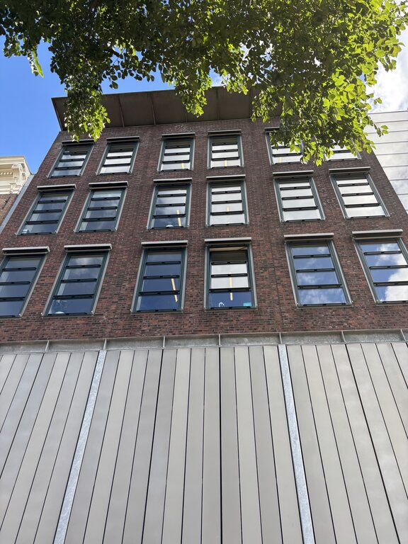Upward view of a brick building in Amsterdam with rows of windows framed by green tree branches against a blue sky.