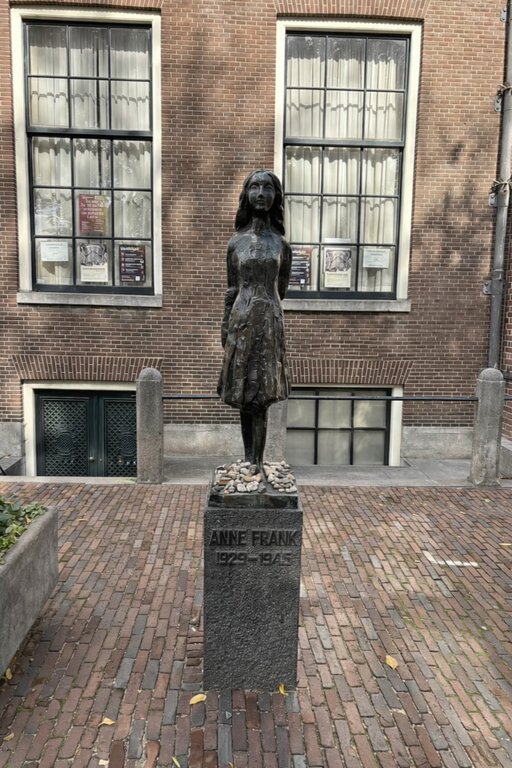 Bronze statue of Anne Frank stands on a pedestal engraved with "ANNE FRANK 1929–1945" in front of a brick building with tall windows in Amsterdam.