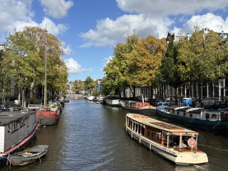 Amsterdam canal lined with houseboats and trees in early autumn with a tour boat moving through the water under a bright sky.