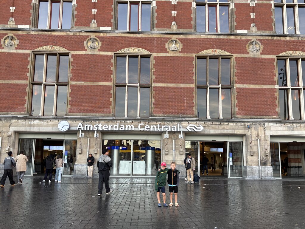 Two children stand in front of the entrance to Amsterdam Centraal station with the sign "Amsterdam Centraal" visible above the doors.