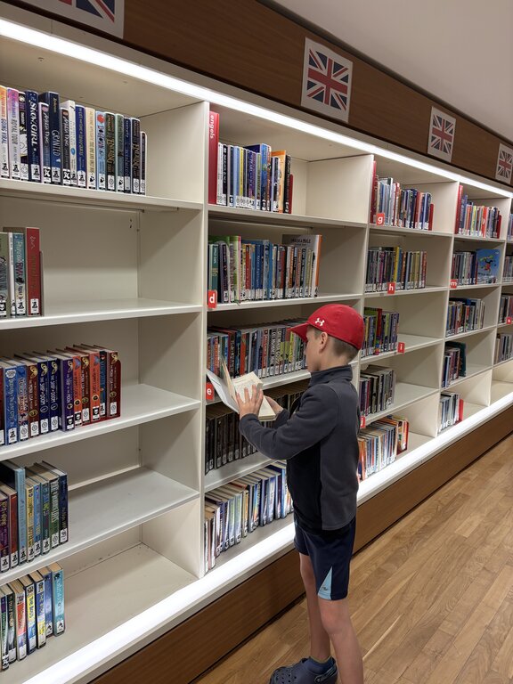 A child wearing a red cap stands in front of a library shelf reading a book with rows of books organized behind him and small UK flags above the shelves.