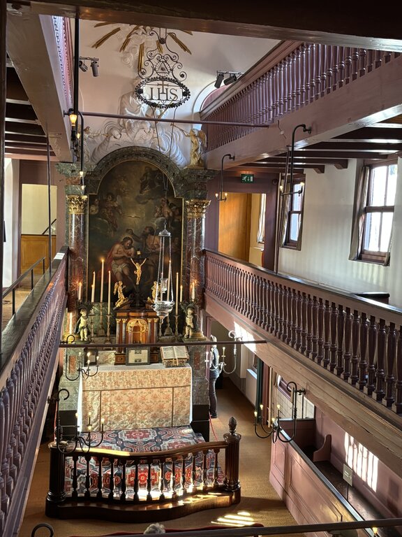 Interior of a hidden church with wooden balconies an ornate altar and religious artwork with candles lit and "IHS" displayed above.