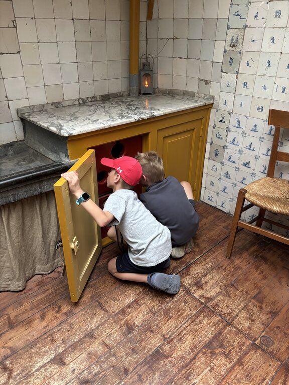 Two children kneel on a wooden floor looking inside a small cabinet in a historic kitchen with tiled walls and a marble countertop.