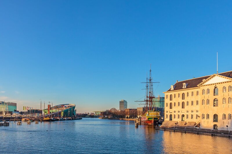 Waterfront view in Amsterdam at sunset with a historic tall ship docked beside a large pale building and modern architecture visible across the water under a clear blue sky.