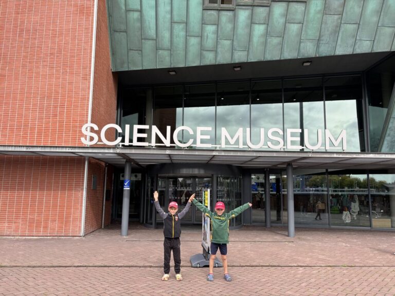 Two children stand with arms raised in front of a building entrance with large letters reading "SCIENCE MUSEUM" above the doors.