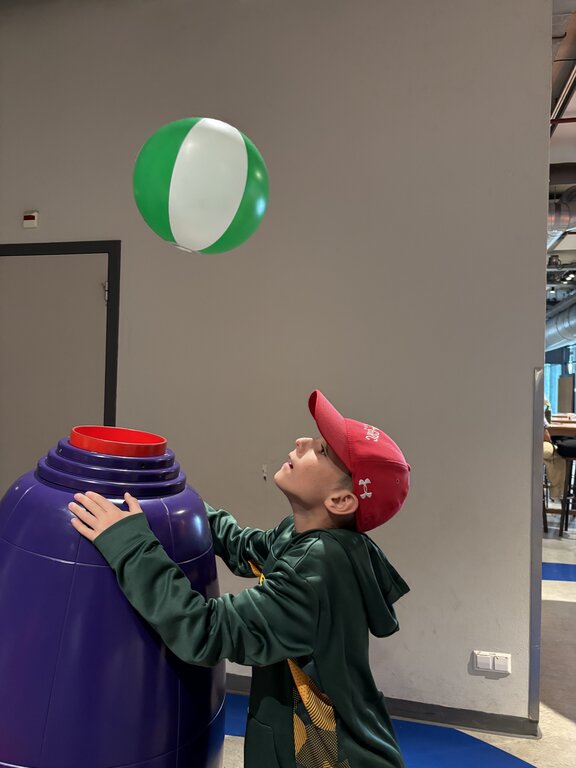 A child wearing a red cap watches a green and white ball float above a purple air blower as part of a hands on science display.