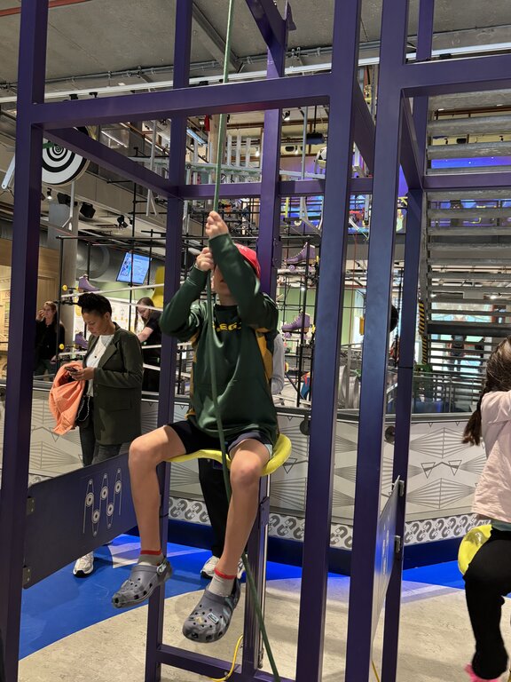A child sits on a small seat pulling a rope as part of an interactive science exhibit inside a museum with colorful structures and other visitors around.
