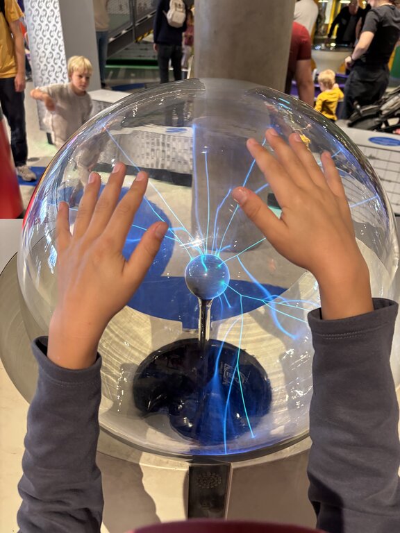 Hands reach toward a plasma globe with blue electric currents glowing inside while other children explore exhibits in the background.