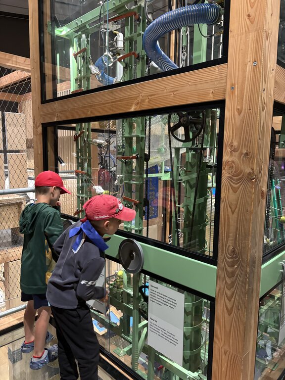 Two children interact with a large glass enclosed mechanical exhibit turning a wheel as gears and tubes move inside at the Nemo Science Museum in Amsterdam.