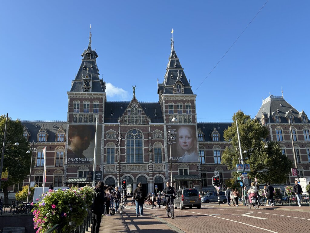 The Rijksmuseum building in Amsterdam with people walking and cycling in front and large banners displayed on the facade.