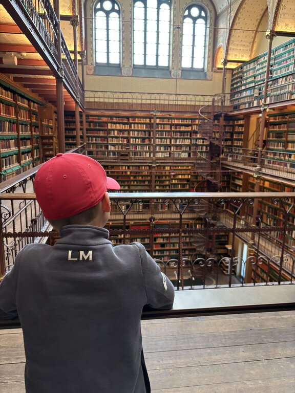 A child looks out over a multi level historic library with tall bookshelves spiral staircase and large arched windows letting in natural light.