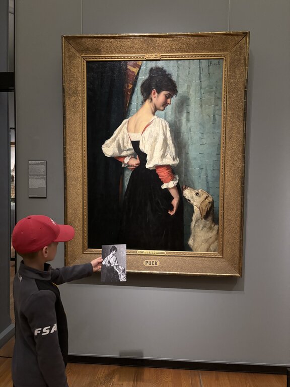 A child in a red cap points to a painting of a woman with a dog with the label "PUCK" visible on the frame at the Rijksmuseum in Amsterdam