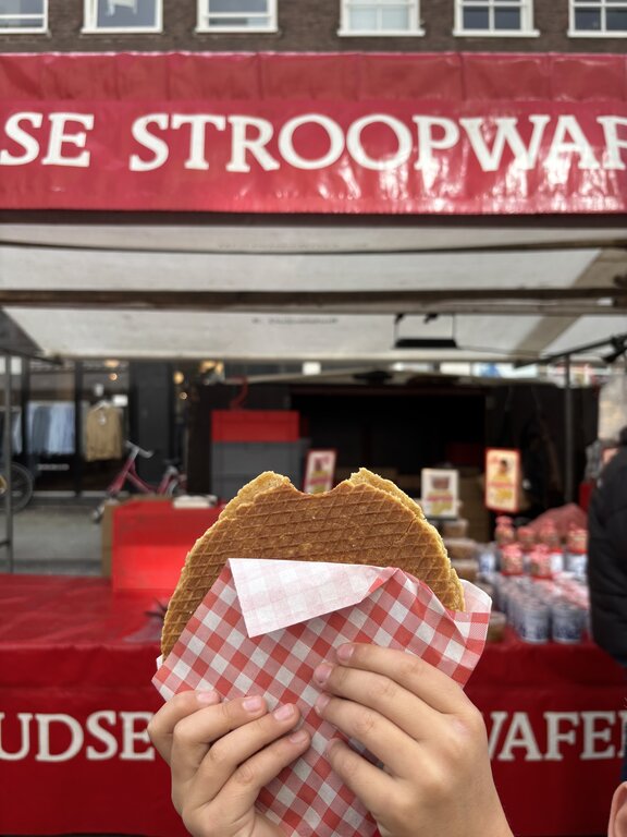 A child holds a freshly made stroopwafel wrapped in red and white paper in front of a market stall with a red sign reading "STROOPWAFEL".
