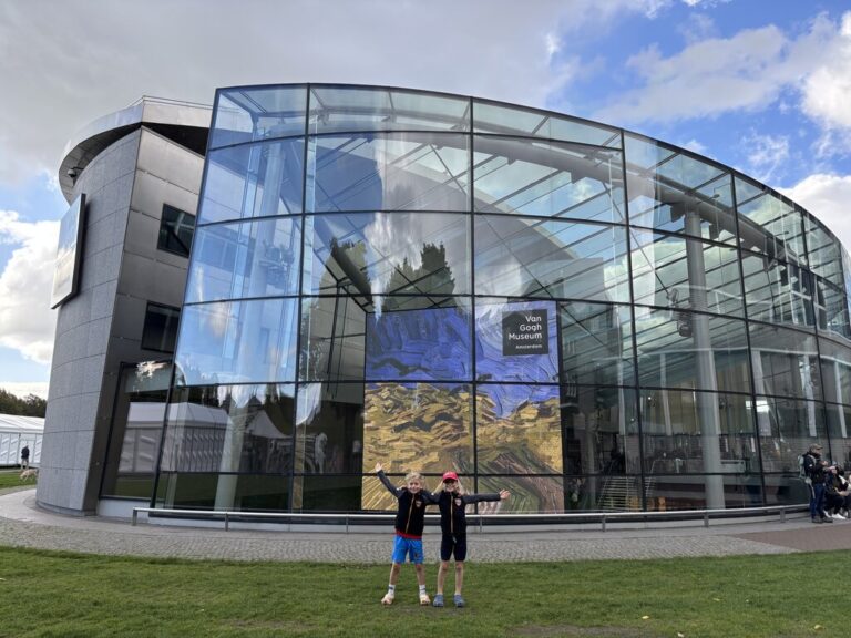 Two kids stand on grass with arms raised in front of the glass exterior of the Van Gogh Museum in Amsterdam with a large artwork display visible through the windows and blue sky above.