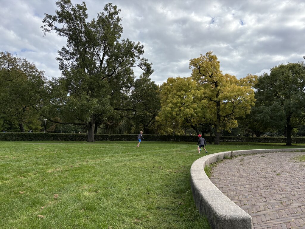 Two children run across a grassy park with large trees and a curved paved path nearby under a cloudy sky in Amsterdam.