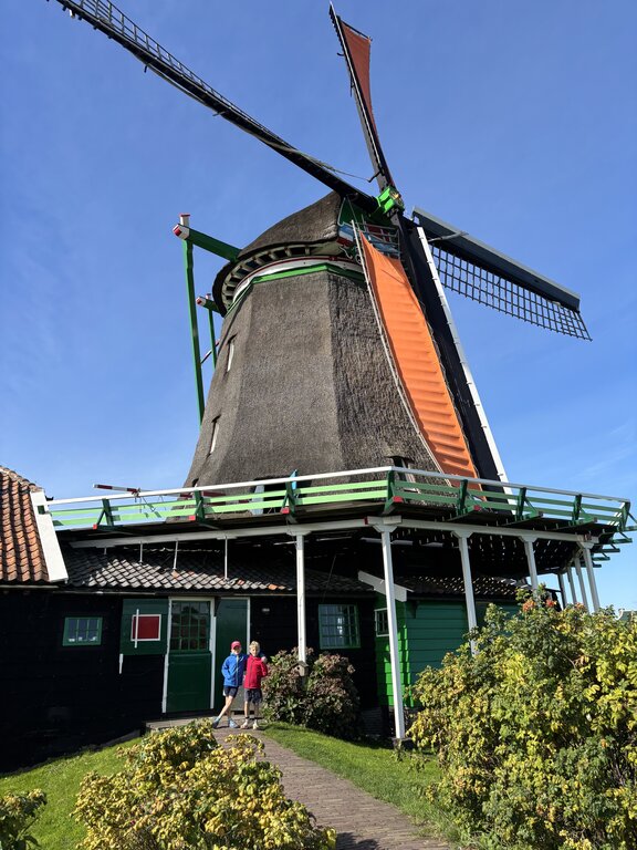 Traditional Dutch windmill with large blades and green accents stands beside a small building with two children walking on the path in front under a clear blue sky.