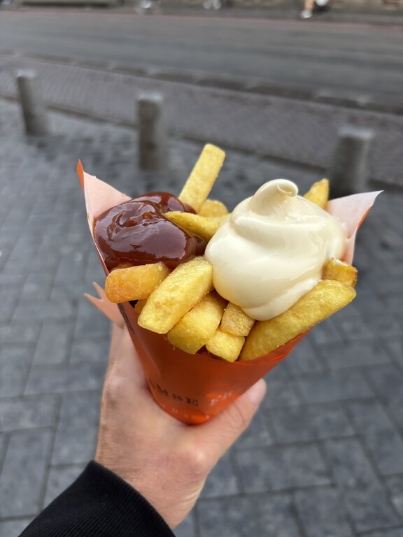 A hand holds a cone of thick cut fries topped with mayonnaise and ketchup while standing on a street in Amsterdam.
