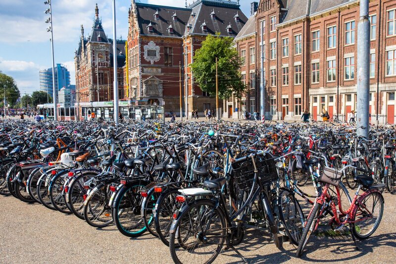 Large crowded bike parking area in Amsterdam filled with hundreds of bicycles in front of historic buildings near a busy street.