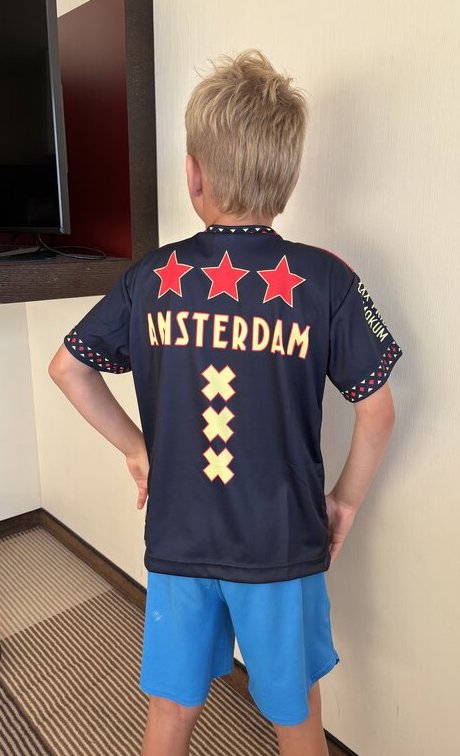 A child stands facing away wearing a dark soccer jersey with three red stars and the word "AMSTERDAM" above three X symbols printed on the back.