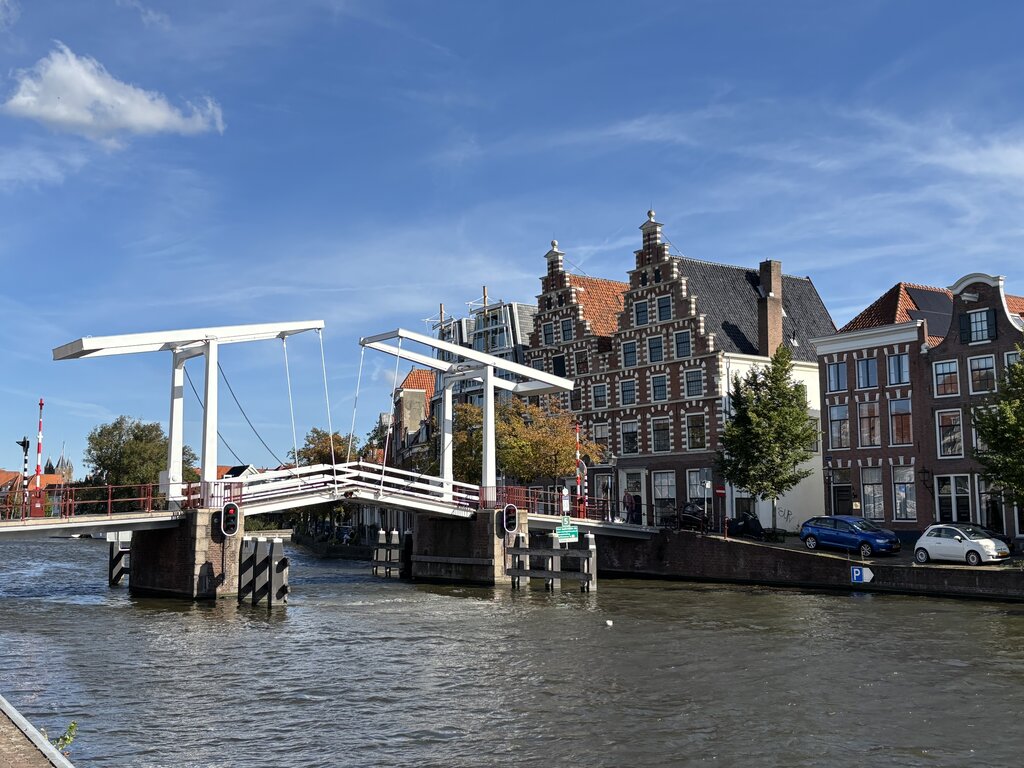 White drawbridge over a canal in Amsterdam with historic buildings behind it and water flowing beneath under a sunny sky.
