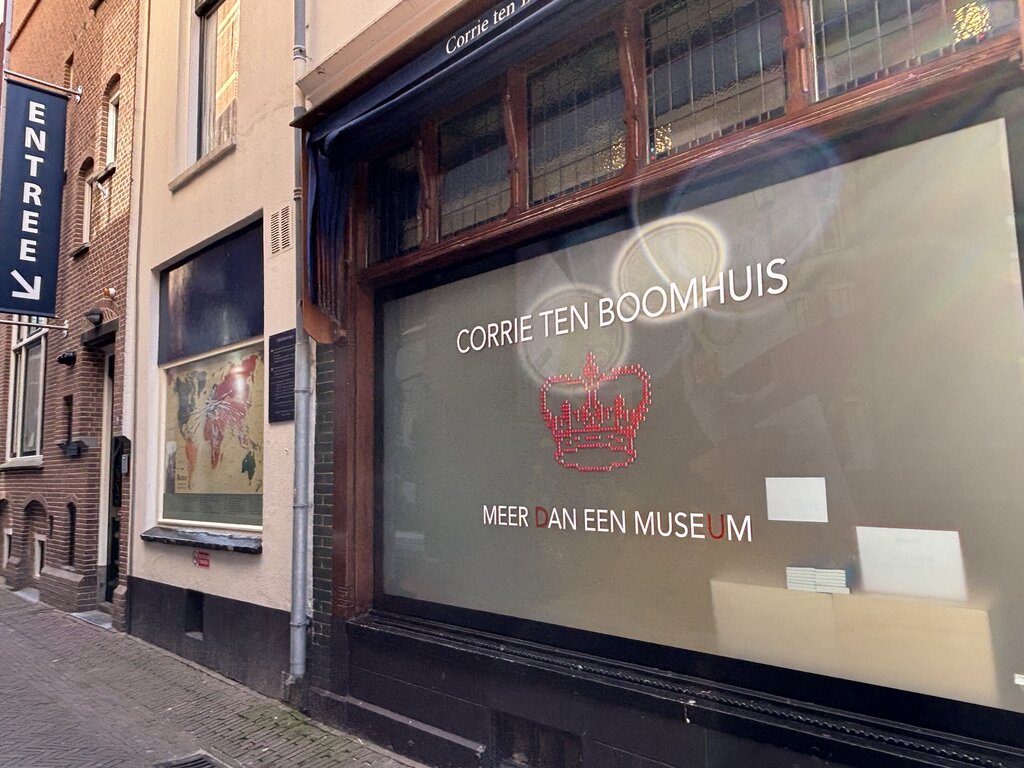 Window display with the text "CORRIE TEN BOOMHUIS" and "MEER DAN EEN MUSEUM" on a glass panel along a narrow street in Amsterdam.