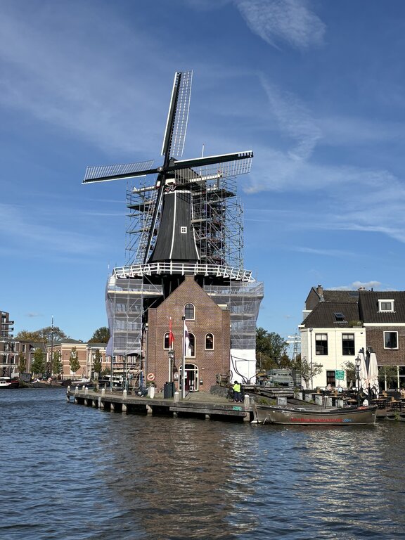 Traditional Dutch windmill surrounded by water and nearby buildings with scaffolding around part of the structure under a blue sky.