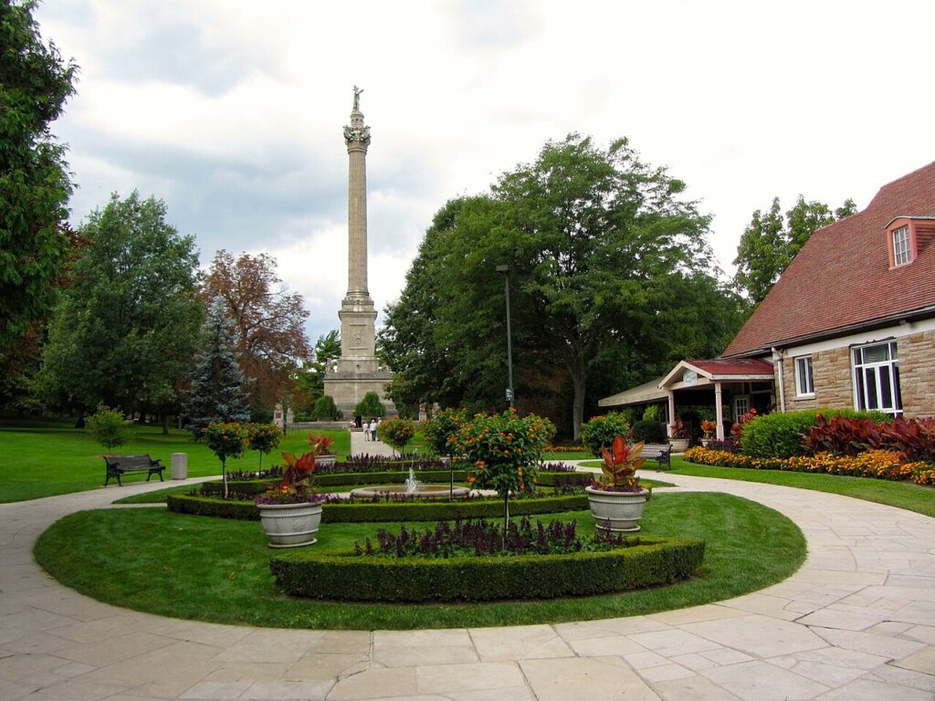 Landscaped park with circular flower beds and a tall stone column monument rising in the center surrounded by trees and a small building with a red roof nearby.