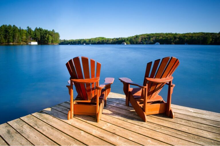 Two wooden Muskoka chairs sit on a dock overlooking a calm lake surrounded by forest under a clear blue sky in Muskoka, Ontario.