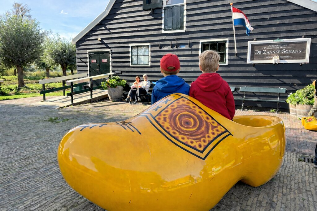 Two children sit inside a giant yellow wooden clog outside a black wooden building with a Dutch flag and sign reading "DE ZAANSE SCHANS".