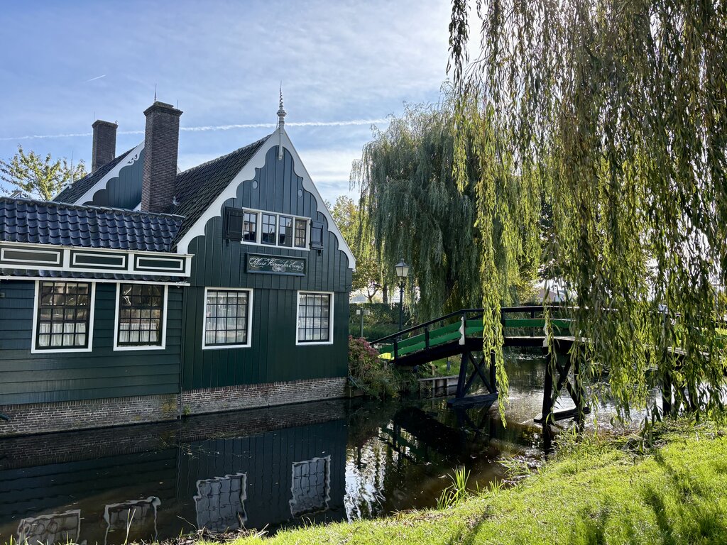 Green wooden house beside a canal with a small bridge and a large tree with hanging branches reflected in the water.