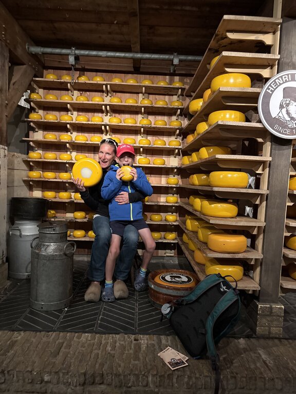 A woman and child sit inside a cheese shop holding large wheels of cheese with shelves stacked full of round yellow cheeses behind them.