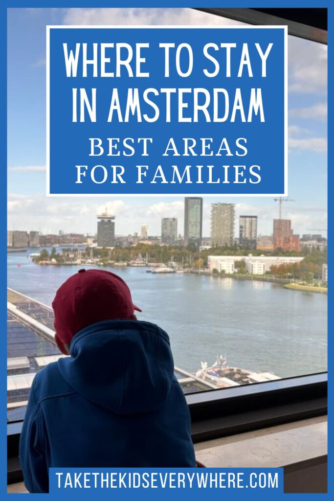 Child in a blue hoodie and red baseball cap looks out a large window over Amsterdam's waterfront and skyline. The image includes the text "WHERE TO STAY IN AMSTERDAM. BEST AREAS FOR FAMILIES." and "TAKETHEKIDSEVERYWHERE.COM".