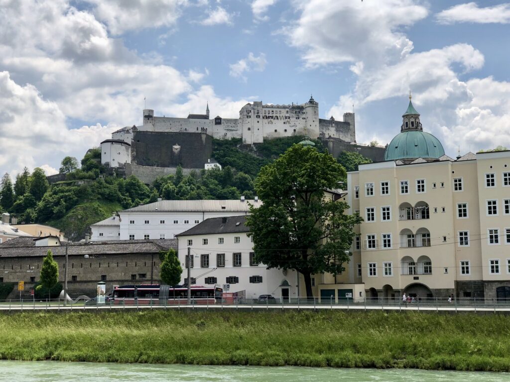 Hohensalzburg Fortress stands above the city skyline while the Salzach River flows through the foreground. Historic buildings and a green domed church complete the classic Salzburg scene.
