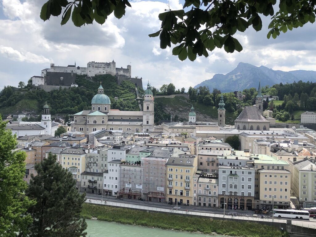 Panoramic view of Salzburg featuring historic rooftops, church domes, and Hohensalzburg Fortress overlooking the city. Mountains in the background add to the dramatic alpine setting.