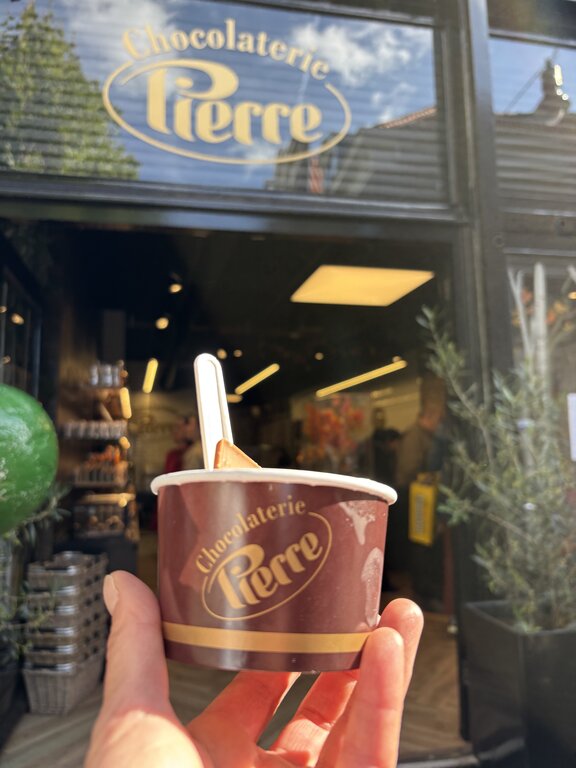 Hand holding a dessert cup outside “Chocolaterie Pierre” in Haarlem, Netherlands. The chocolate shop entrance glows warmly in the background with the store logo visible above the doorway.
