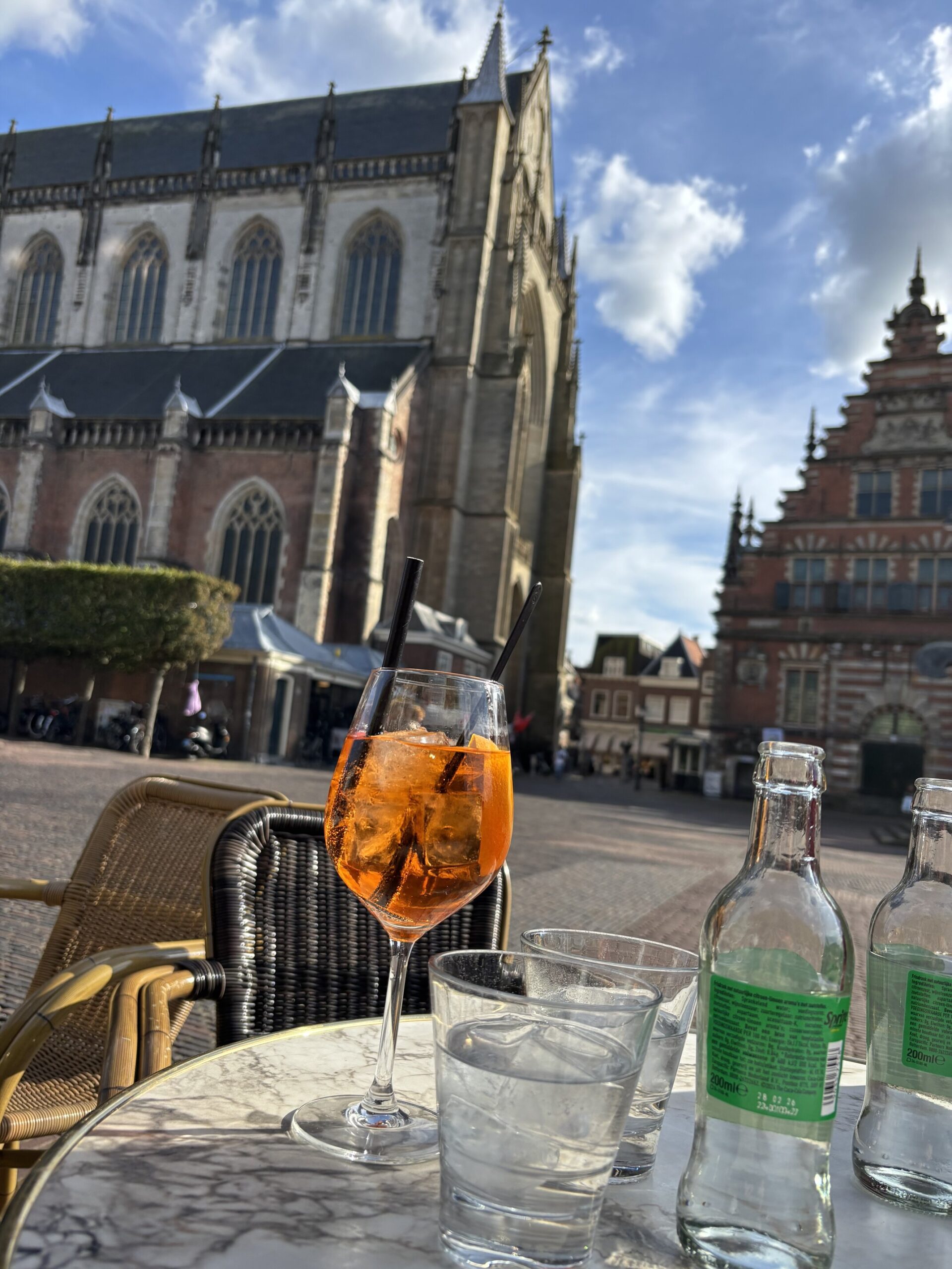 Orange cocktail on an outdoor café table in Haarlem, Netherlands, with water glasses and bottles in the foreground. The Grote Kerk church and historic Dutch buildings stand behind the terrace beneath a bright blue sky with scattered clouds.