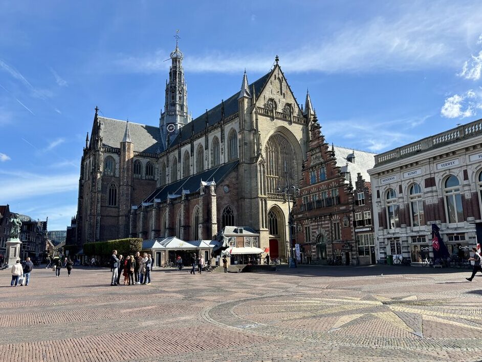 Large historic church in a wide public square in Haarlem with people walking across the open cobblestone plaza under a clear sky.
