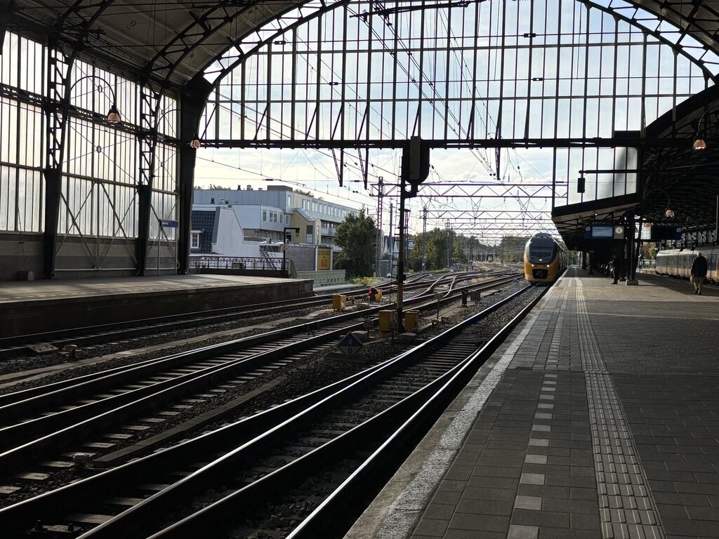 Large covered train station in Haarlem, Netherlands, with multiple railway tracks leading toward a yellow train in the distance. Sunlight filters through the tall glass and steel roof structure above the empty platform.