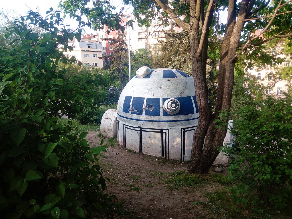R2 D2 inspired sculpture partially hidden among trees and shrubs in a small city park. The unexpected Star Wars themed artwork is one of the more quirky and unusual things to see in Prague.
