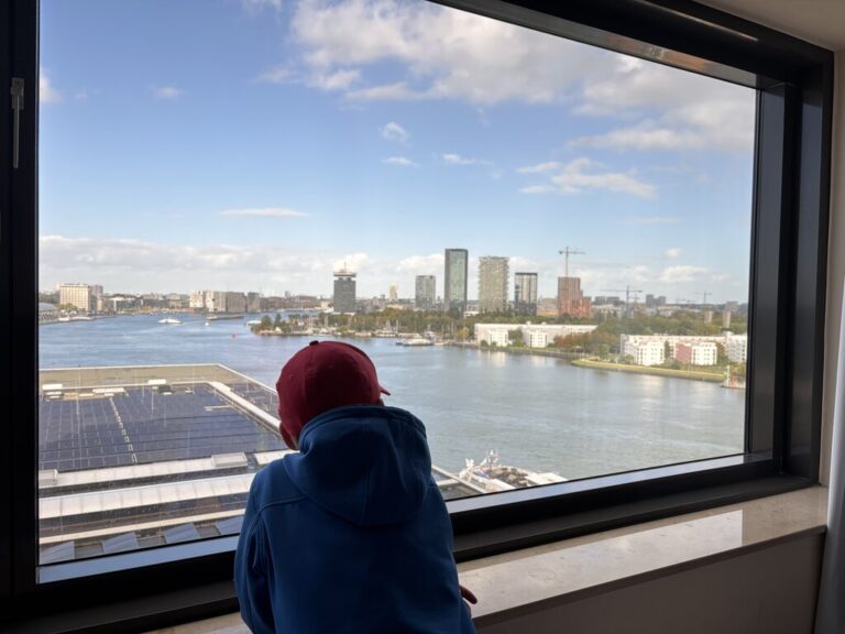 Child looking out the window at the waterfront and city skyline from a room at the Mövenpick Hotel Amsterdam City Centre.
