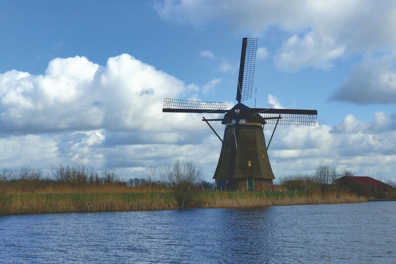 A traditional Dutch windmill rises above the water at Kinderdijk in the Netherlands beneath a sky filled with large drifting clouds. Tall grasses and reeds line the shoreline in front of the mill.