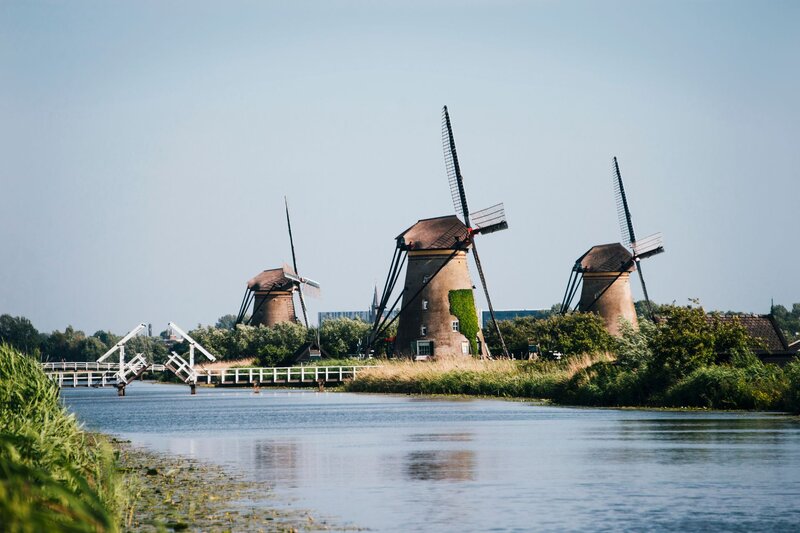 Three historic windmills stand along a wide canal at Kinderdijk in the Netherlands with a white drawbridge visible in the distance. Calm water reflects the mills and the surrounding green landscape.