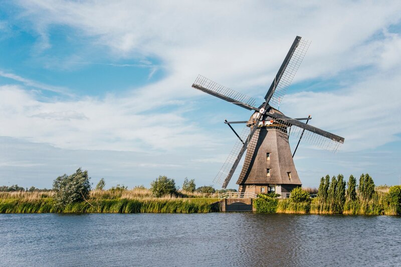 A single traditional windmill stands on the riverbank at Kinderdijk in the Netherlands beneath a soft blue sky streaked with thin clouds. Trees and tall grasses surround the mill beside the water.